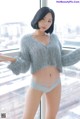 A woman in a gray sweater and panties posing by a window.